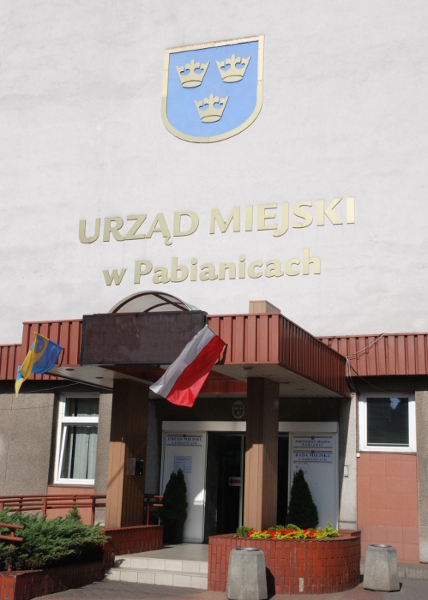 budynek urzędu, zadaszone wejście z powiewającymi na wietrze flagami, nad nim złoty napis Urząd Miejski w Pabianicach i herb miasta Pabianice